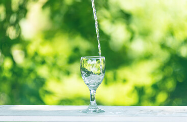 Water flows into a glass placed on a wooden bar,over sunlight and natural green background.blur shadow.
