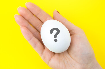 White egg with question mark  in woman hand on the yellow background. Pregnancy and baby concept. Who is? Copy space. Minimalism, original and creative photo. Beautiful wallpaper. Easter holidays.