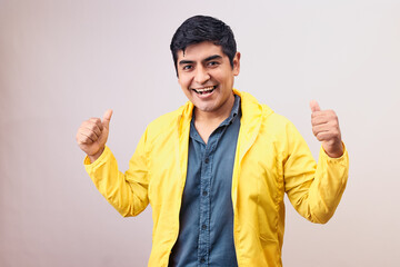 Hombre joven y feliz levanta pulgares. Modelo aislado en fondo blanco con casaca amarilla