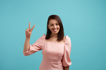 Joyful beautiful woman in pink dress shows peace sign and welcomes everyone smiles positively, makes victory gesture, isolated over blue background. People expression concept.