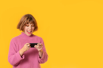 A girl stands in front of a yellow background and She plays with her cell phone. She is very happy. Gamer girl is busy with attention a lot.
