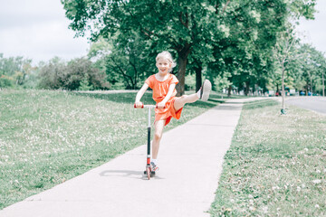 Cute funny Caucasian girl child in red orange romper riding scooter on street road park outdoor. Summer fun eco sport activity for kids children. Authentic real candid childhood lifestyle