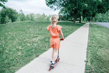 Young girl child in red orange romper riding scooter on street road park outdoor. Summer fun eco sport activity for kids children. View from back behind. Authentic real candid childhood lifestyle © anoushkatoronto