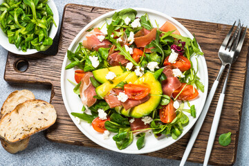 Green salad with avocado, salad leaves, jamon serrano and tomatoes. Healthy diet lunch. Top view at light stone table.
