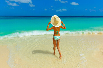 Naklejka na ściany i meble Girl in bikini having fun at the beach with beach hat standing on the shore looking at horizon, interracial, black 