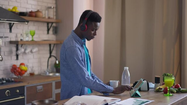 Side view of young concentrated man in headphones drinking morning coffee surfing Internet checking e-mail on tablet standing at kitchen countertop. Focused African American millennial in home office