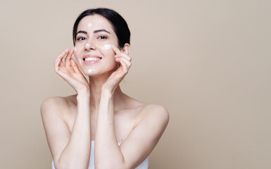 Close up beauty portrait of a laughing beautiful half naked woman applying face cream over beige background with copy space