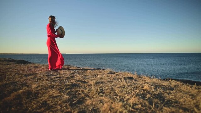 An attractive young Caucasian woman in a red dress with long hair stands with a straw hat in her hands on a rocky seashore with closed eyes in windy weather. Shallow depth of field