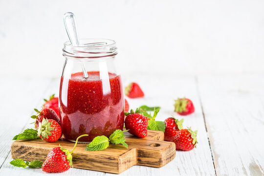 Strawberry jam in the glass jar at white table with fresh strawberries.