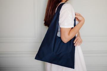 young girl in a white dress holds a blue cotton shopper bag on her shoulder in the interior. the...