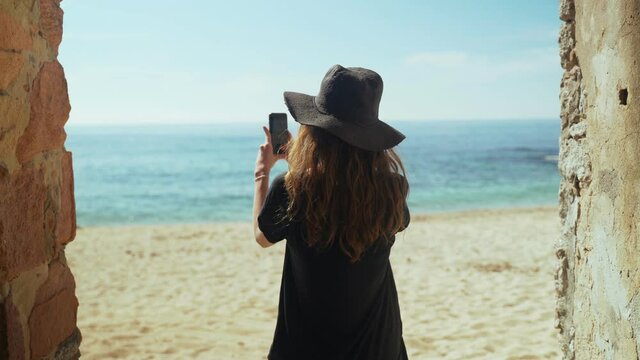 Young woman in wide black pants and a hat stands in an old lane overlooking the beach, enjoys the sea view and making photo with smartphone. Sunny day on the beach. Holidays. Summer chill. White sand.
