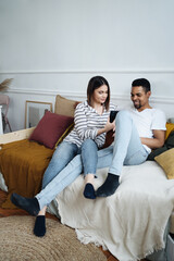 Happy multiethnic couple sit on couch at home watching funny video on smartphone, smiling loving international family husband and wife relax on sofa in living room using mobile together