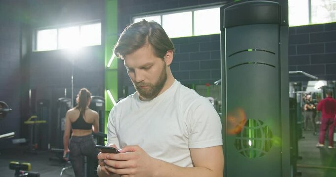 Strong athlete bearded man is standing at the gym and resting after hard training. Guy is looking at smartphone screen while holding gadget in hand. Stock video