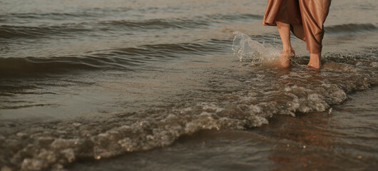 female legs: a woman in a gold dress walks along the river bank, waves at sunset