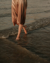 female legs: a woman in a gold dress walks along the river bank, waves at sunset