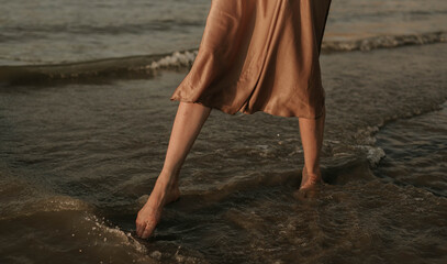 female legs: a woman in a gold dress walks along the river bank, waves at sunset