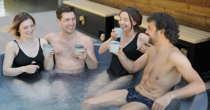 Company of friends bathing in outdoor jacuzzi or hot vat at the luxury spa. People relax and have fun drinking hot drinks in wellness center