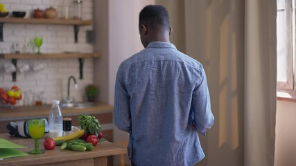 Live camera follows young confident African American serious man with business graph walking in kitchen to table opening laptop and sitting down on chair. Start of work time in home office. New normal