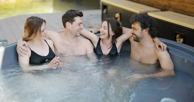 Company of friends bathing in outdoor jacuzzi or hot vat near the spa and lake. Couples have fun and relax in wellness center