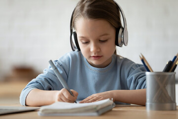 Close up of smart small schoolgirl in headphones write prepare homework alone at home. Little Caucasian 7s girl child in earphones handwrite study online distant, home assignment. Education concept.