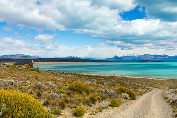 paisagem com uma estrada de terra cercada por grama e flores com a casa de madeira à esquerda e o fundo o lindo lago azul com montanhas nevadas em um lindo dia de sol
