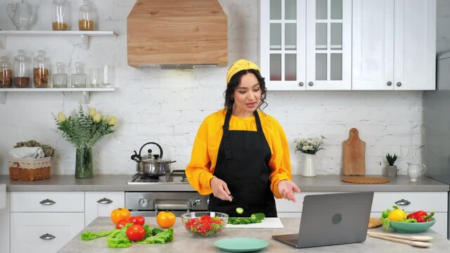 Smiling woman housewife in black apron tells chef teacher slices cucumber, study online video conference call webcam chat laptop computer, distance education culinary course lesson in home kitchen