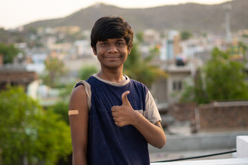 Portrait of an Indian teen giving thumbs up after getting vaccinated and showing bandage on his arm and giving thumbs up