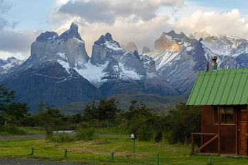 Papier Peint photo autocollant Cuernos del Paine Chalet en bois avec toit vert avec pelouse verte et buissons autour et en arrière-plan le magnifique paysage des montagnes Cuernos del Paine