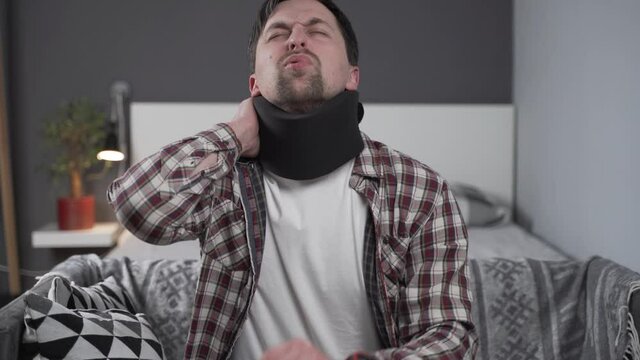 Guy wearing neck brace to support neck at home on couch. Caucasian man with neck injury uses collar support to relieve pain. Male in cervical brace relieves tension and pain in muscles of spinal