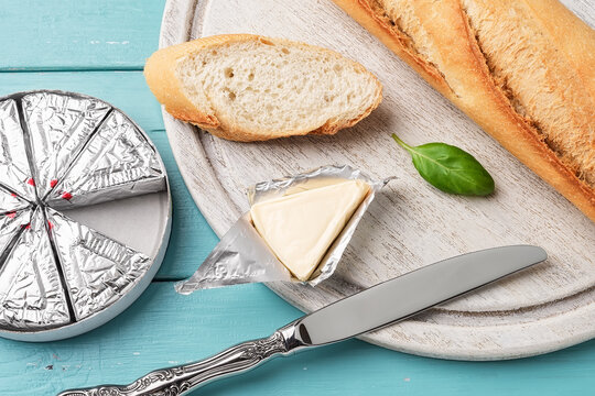 Foil wrapped processed cream cheese, table knife and slice of bread on a cutting board over blue wooden table. Small triangular pieces of soft cheese in an aluminium foil. Tasty sandwich ingredient.