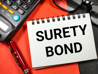 Business concept.Text SURETY BOND with pen,calculator and glasses on red and black background.