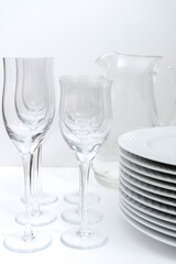 Set of stacked white plates, crystal stemware and a glass jug on a white table. Portrait Photography.