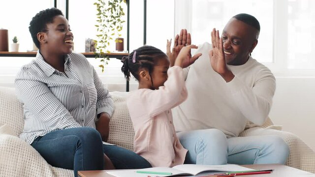 Happy african family afro american parents helping black little girl schoolgirl child draw picture homework sitting on couch at table baby giving high five to mom and dad successful teamwork gesture
