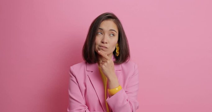 Stylish elegant woman in business costume holds chin looks around with thoughtful expression tries to decide then smiles gladfully has dark hair isolated over pink background. Let me think concept
