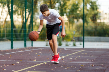Obraz premium A cute boy in a white t-shirt plays basketball on a city playground. Active teen enjoying an outdoor game with an orange ball. Hobby, active lifestyle, sport for kids.