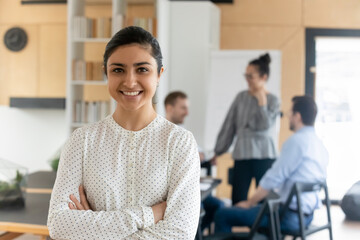 Head shot portrait smiling confident Indian businesswoman with arms crossed standing in modern office with colleagues on background, successful happy employee entrepreneur executive looking at camera - Powered by Adobe