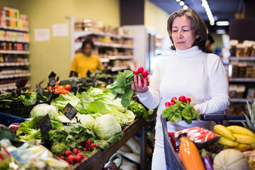 Obraz premium Portrait of positive elderly woman buying fresh organic vegetables in supermarket, choosing red radish