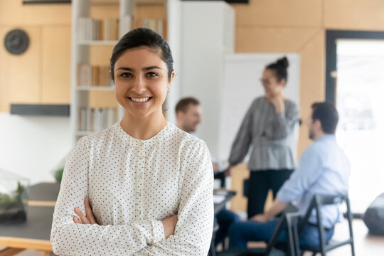 Head shot portrait smiling confident Indian businesswoman with arms crossed standing in modern office with colleagues on background, successful happy employee entrepreneur executive looking at camera