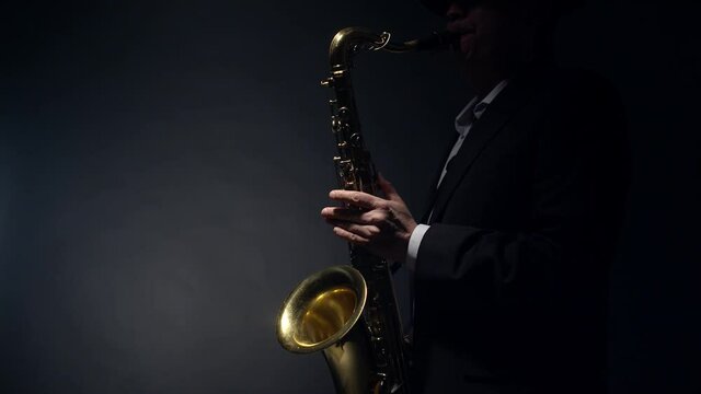 Saxophonist plays a shiny gold saxophone by blowing into the mouthpiece and pressing the flaps with fingers in studio on gray background, profile view. Musician man plays a saxophone.
