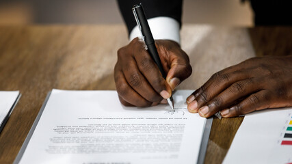 Close up and selective focus on African black formal professional business male hands holding pen, signing in white paper form or application to confirm and deal contract agreement on table.