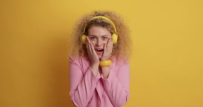 Surprised cheerful curly haired young woman says wow opens mouth feels impressed cannot believe in amazing news wears stereo headphones on ears elegant pink jacket isolated over yellow background.