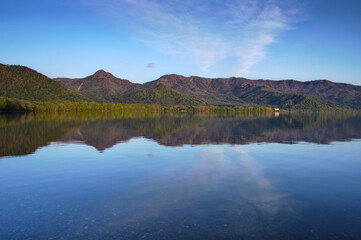 Fototapeta na wymiar 静水の湖面に映る湖畔の森と山々と空。
