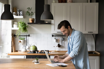 Happy young man in glasses standing in kitchen, calculating domestic utility bills, paying for services online using laptop e-banking application, managing household payments or planning investment.