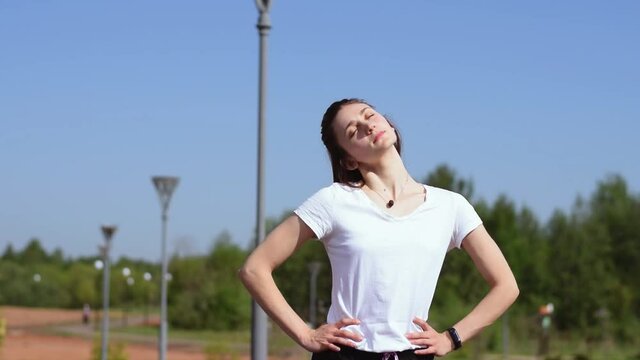 Athletic girl in a white T-shirt warms up the muscles of the neck, shoulders, arms and chest before training. Sports healthy lifestyle. Outdoor training. Slow motion.