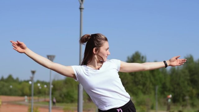 Athletic girl in a white T-shirt warms up the muscles of the neck,shoulders,arms and chest before training.Body tilts to the sides.Sports healthy lifestyle.Outdoor training.Slow motion.
