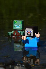 Fototapeta premium Minecraft LEGO figure of Steve chased by drowned zombie hostile mob figure in shallow waters, daylight sunshine.