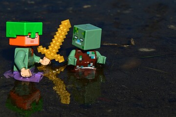 Fototapeta premium Minecraft LEGO figure of Alex with golden sword and tortoise shell helmet attacking suprised drowned zombie mob in shallow waters. Daylight spring sunshine.