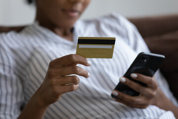 Close up millennial african woman holding cellphone and banking card in hands, enjoying shopping in mobile application, easy modern technology usage, secure internet payments, consumerism concept.