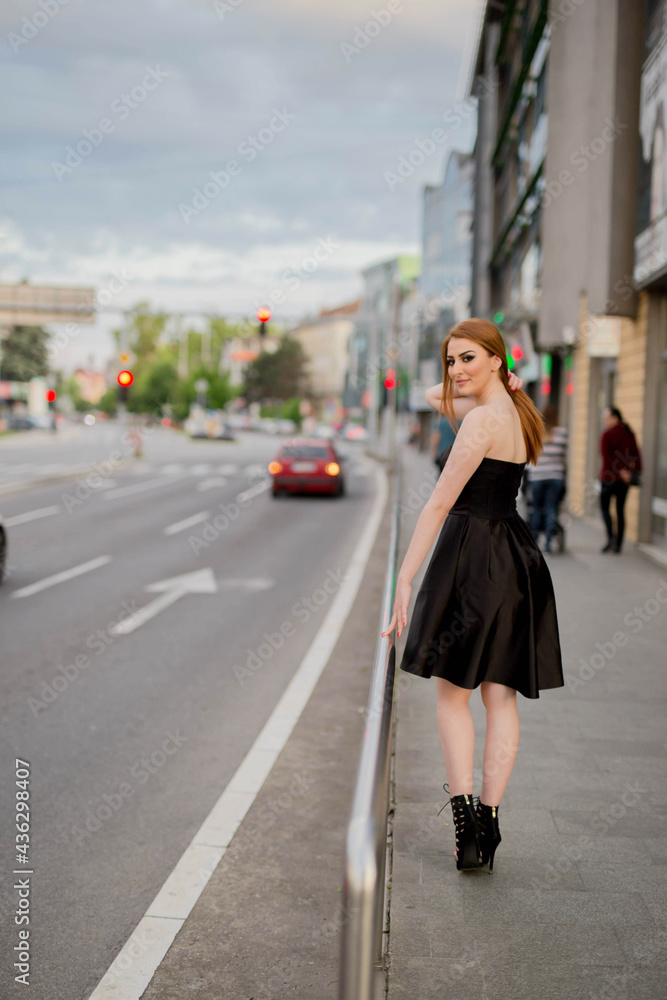 Poster Young Caucasian female with beautiful makeup wearing an elegant black dress walking on a street