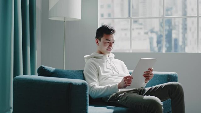 Smiling Asian guy in hoodie watches videos on new tablet